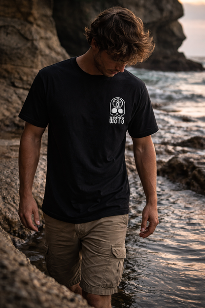 Man wearing a black t-shirt with a logo standing on a rocky beach.