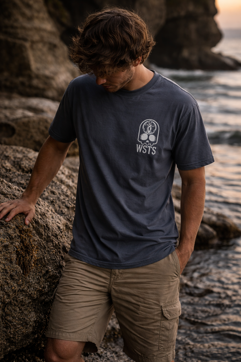 Person wearing a dark blue t-shirt with a logo standing on a rocky shore.